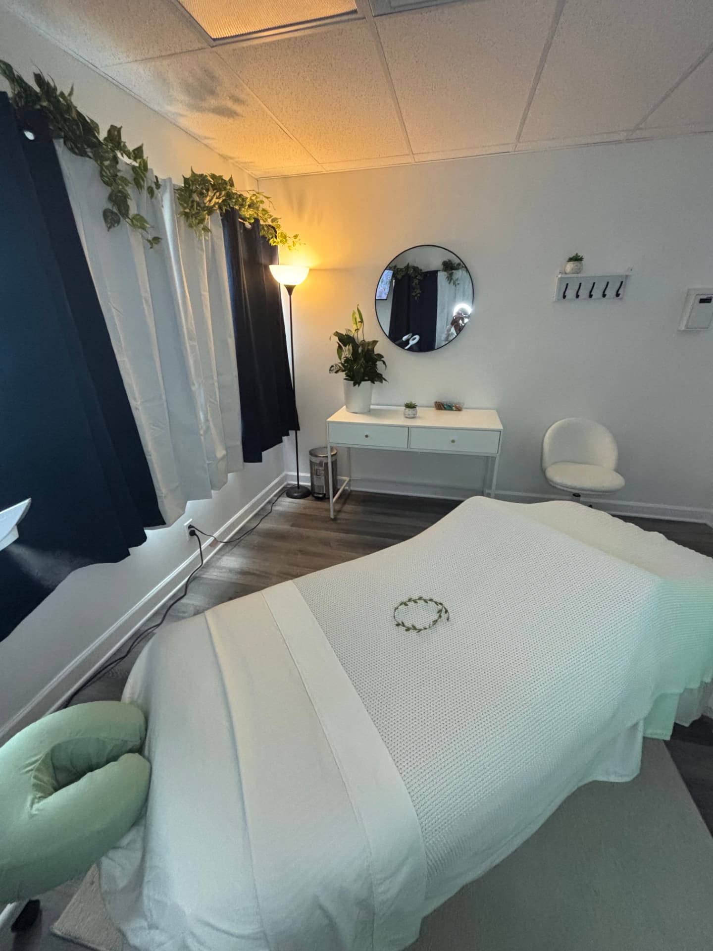 The Vital Bloom treatment room in Lauderdale Lakes, with a dressed massage table, trailing greenery, plant, and warm lamp light.